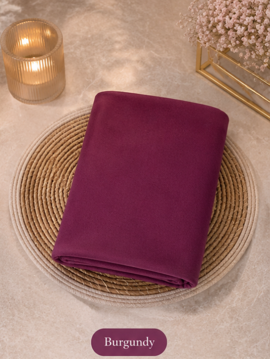 Folded Deep burgundy liquid chiffon hijab fabric on a woven mat with a candle and decorative elements in the background.
