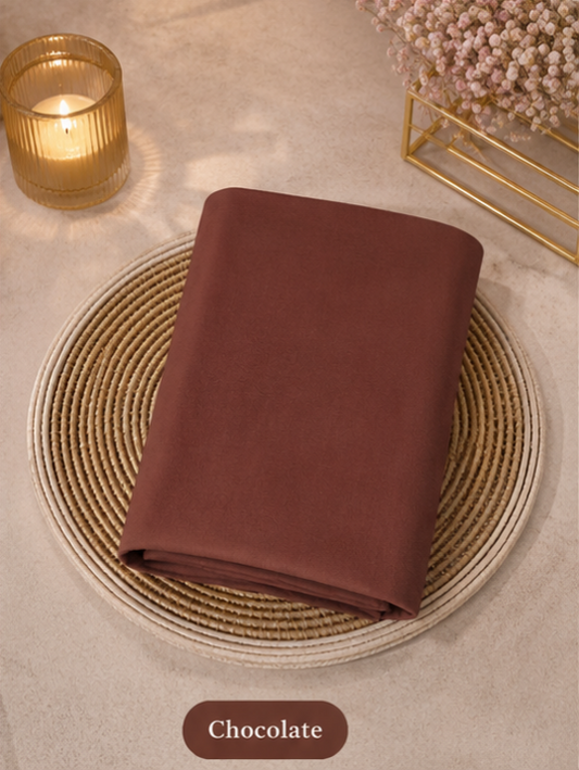 Folded chocolate-colored Liquid Chiffon Hijab fabric on a woven placemat with a candle and decorative elements in the background.