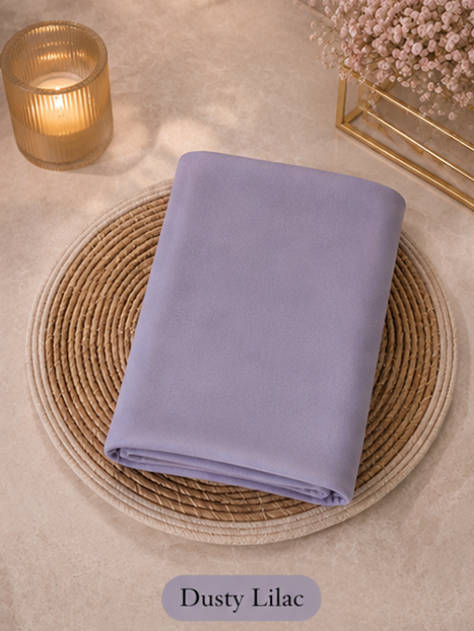 Folded dusty lilac Liquid Chiffon Hijab fabric on a woven mat with a candle and flowers in the background
