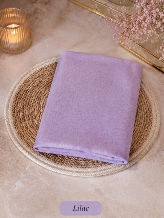 Folded lilac Liquid Chiffon Hijab fabric on a woven mat with a candle and flowers in the background