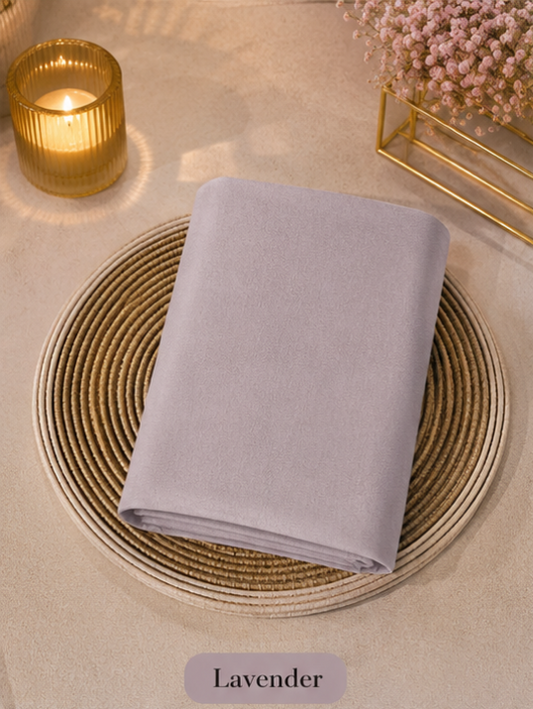 Folded lavender Liquid Chiffon Hijab fabric on a woven placemat with a candle and flowers in the background.