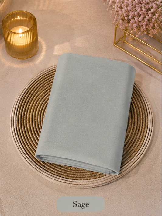 Sage-colored folded Liquid Chiffon Hijab fabric on a woven mat with a candle and flowers in the background