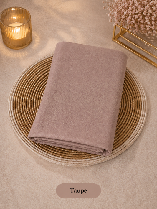 Taupe folded Liquid Chiffon Hijab on a woven placemat with a candle and decorative elements in the background.