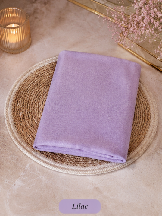 Folded lilac Liquid Chiffon Hijab fabric on a woven mat with a candle and flowers in the background